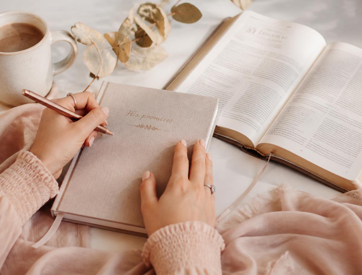 A woman writing in a linen journal beside an open Bible in soft morning light